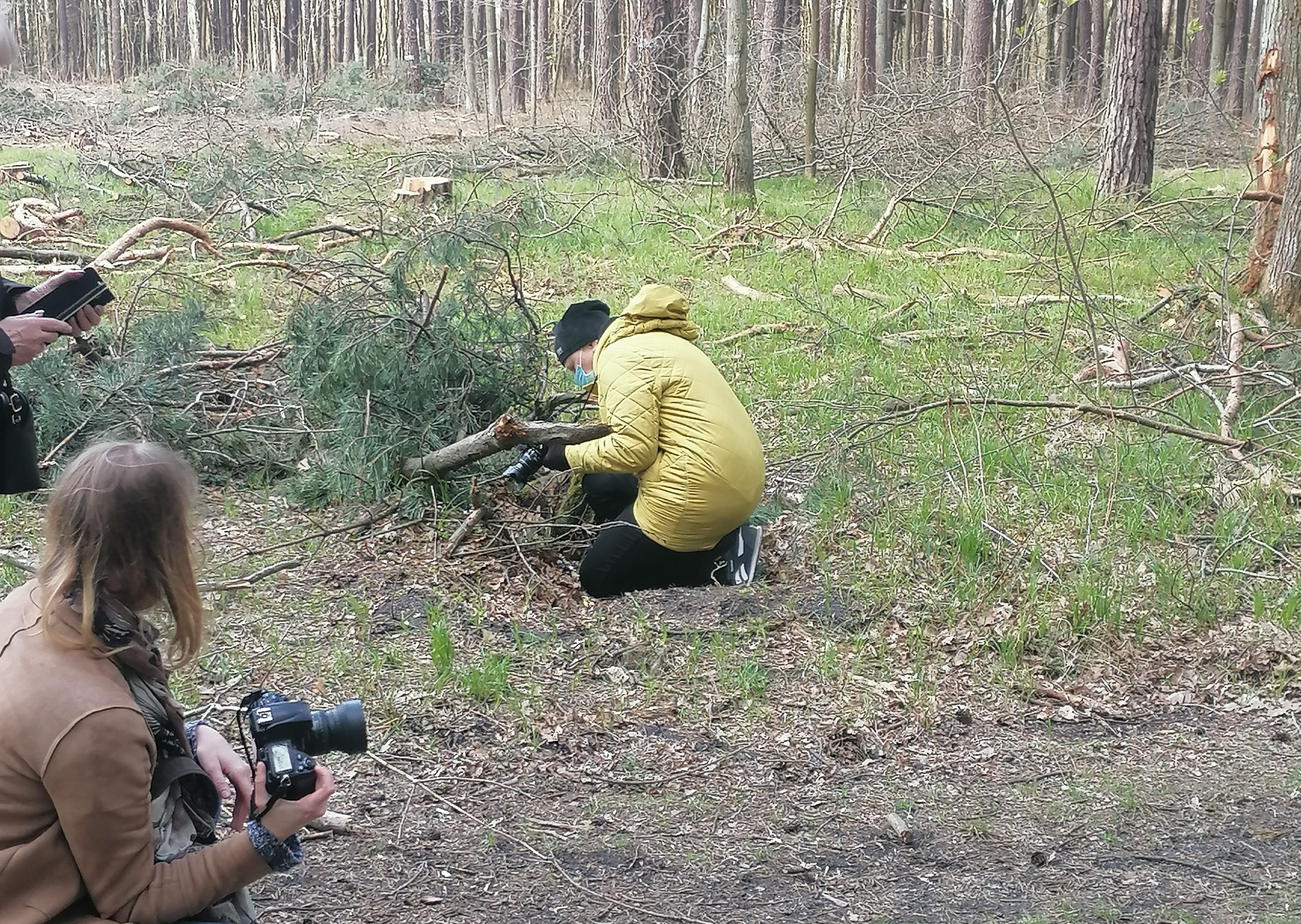 Plenerowe warsztaty fotograficzne Klub Seniora Platyna - zdjęcie pokazuje las i uczestników fotografujących gałęzie na leśnej polanie. Nad procesem czuwa instruktorka.