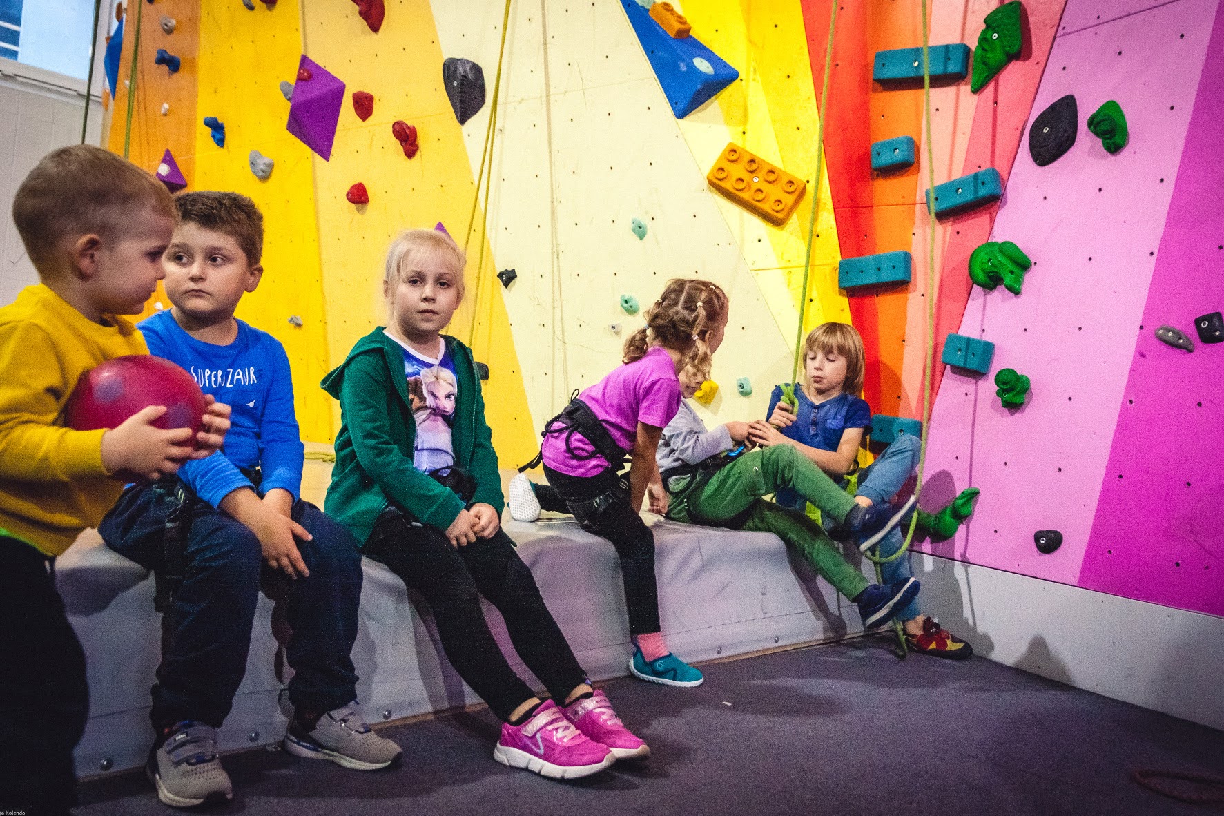Zdjęcie z zajęć sportowych przedstawia dzieci siedzące pod ścianką wspinaczkową
