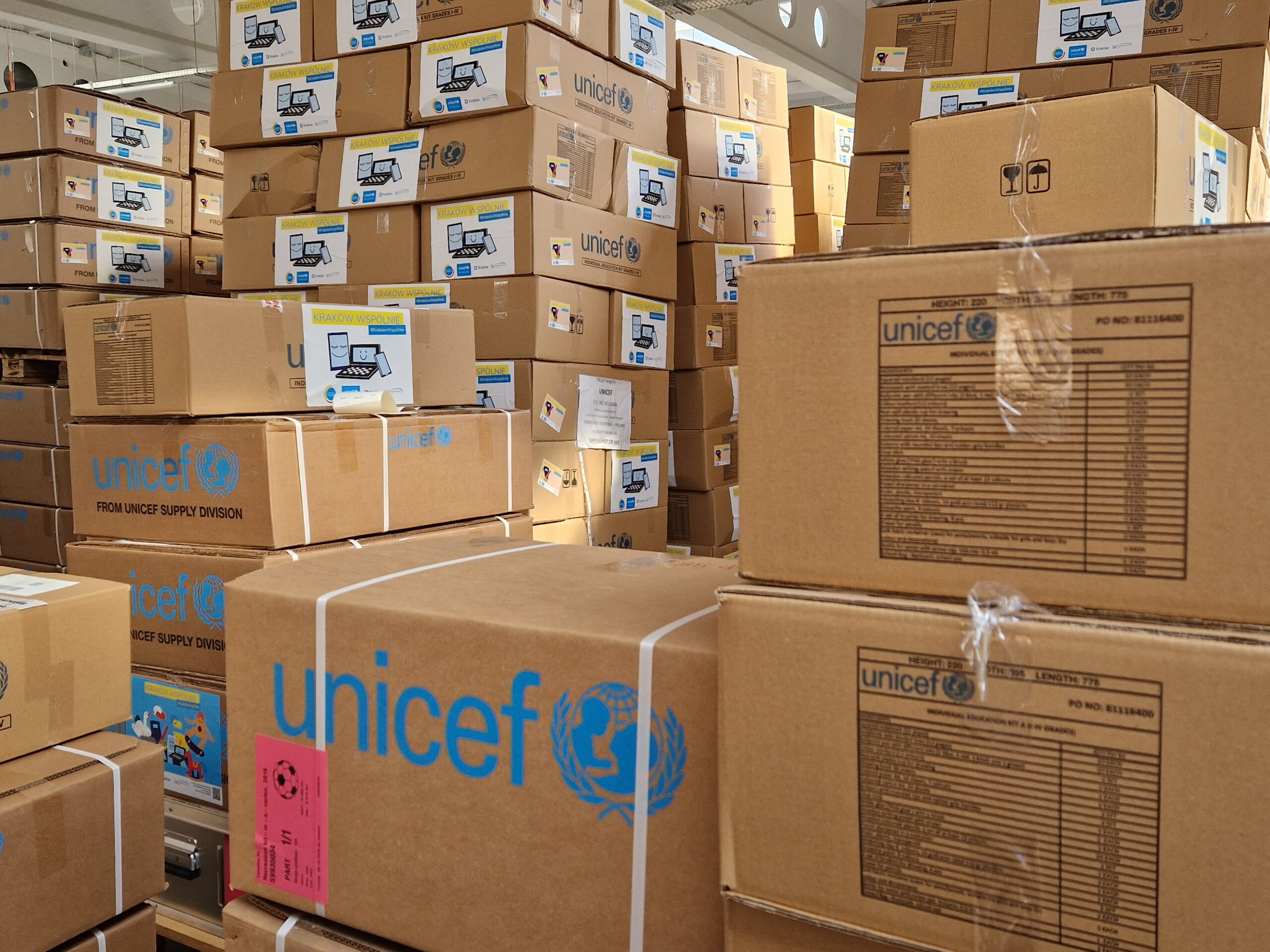Zdjęcie przedstawia magazyn zapełniony kartonami z przyborami. Na kartonach widnieje lolo Kraków Wspólnie oaz UNICEF.