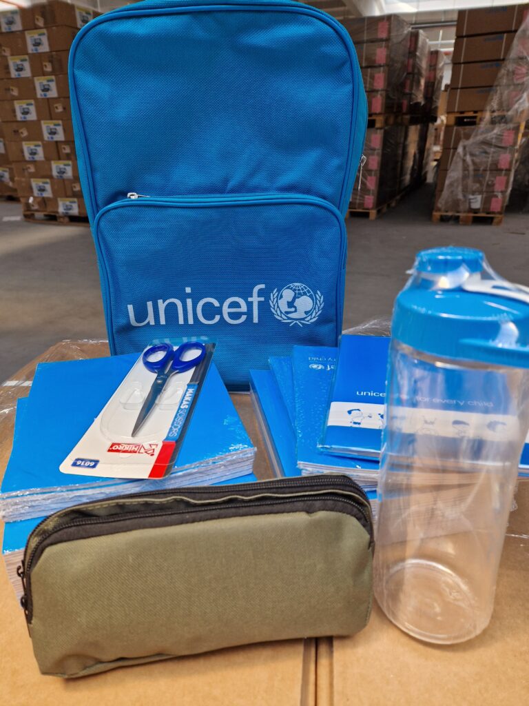 Zdjęcie przedstawia przykładową wyprawkę dla uczniów: plecak z logo UNICEF, piórnik, bidon na wodę, zeszyty, przybory szkolne.